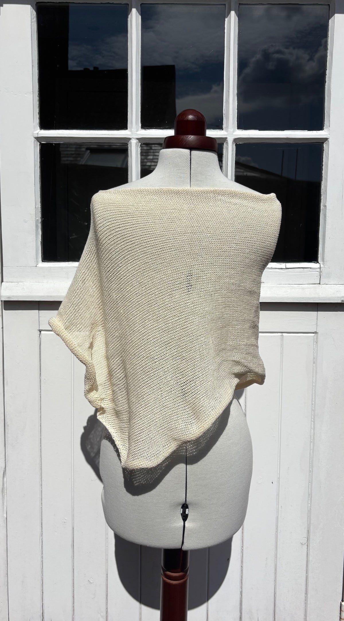 A cream-colored, textured knit poncho draped over a white dress form mannequin, set against a white wooden paneled door.