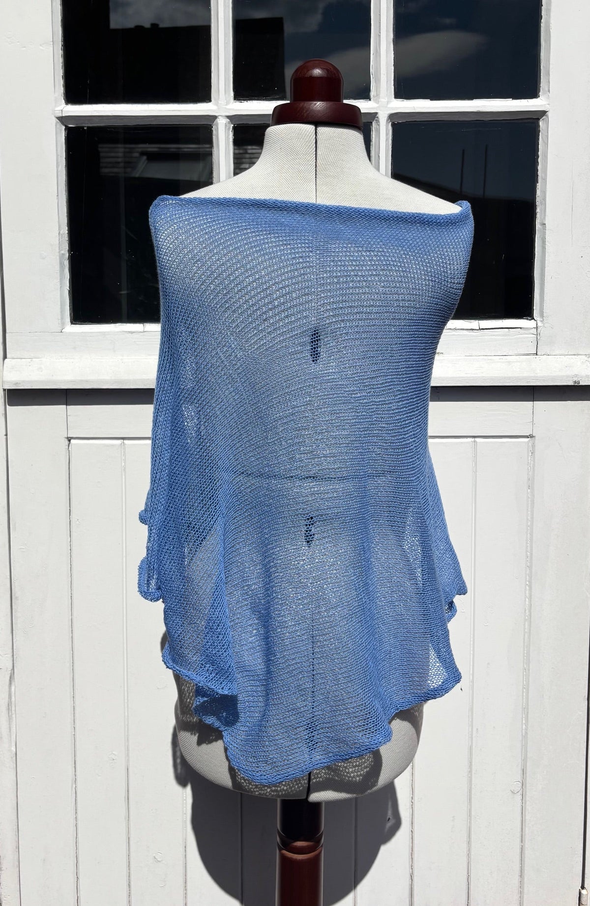 A light blue open-knit shawl or wrap draped over a dress form mannequin, set against a white paneled door with window panes.