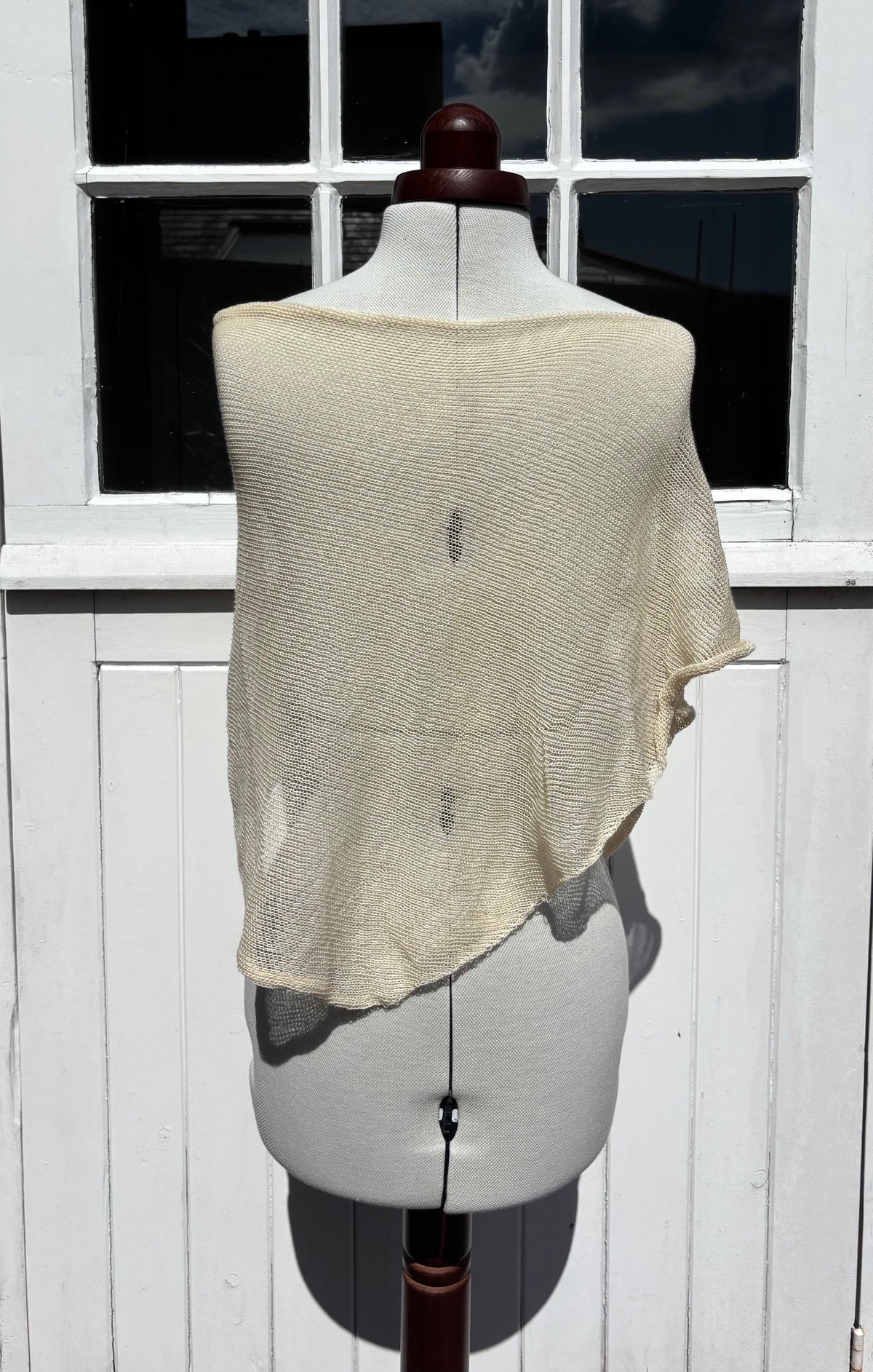 A cream-colored, semi-sheer open-knit shawl or wrap draped over a white dress form mannequin, set against a white wooden paneled wall with a window.