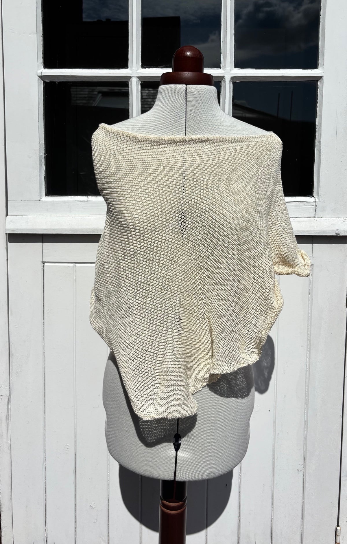 A cream-colored, textured knit poncho draped over a gray dress form mannequin, set against a white wooden paneled door.