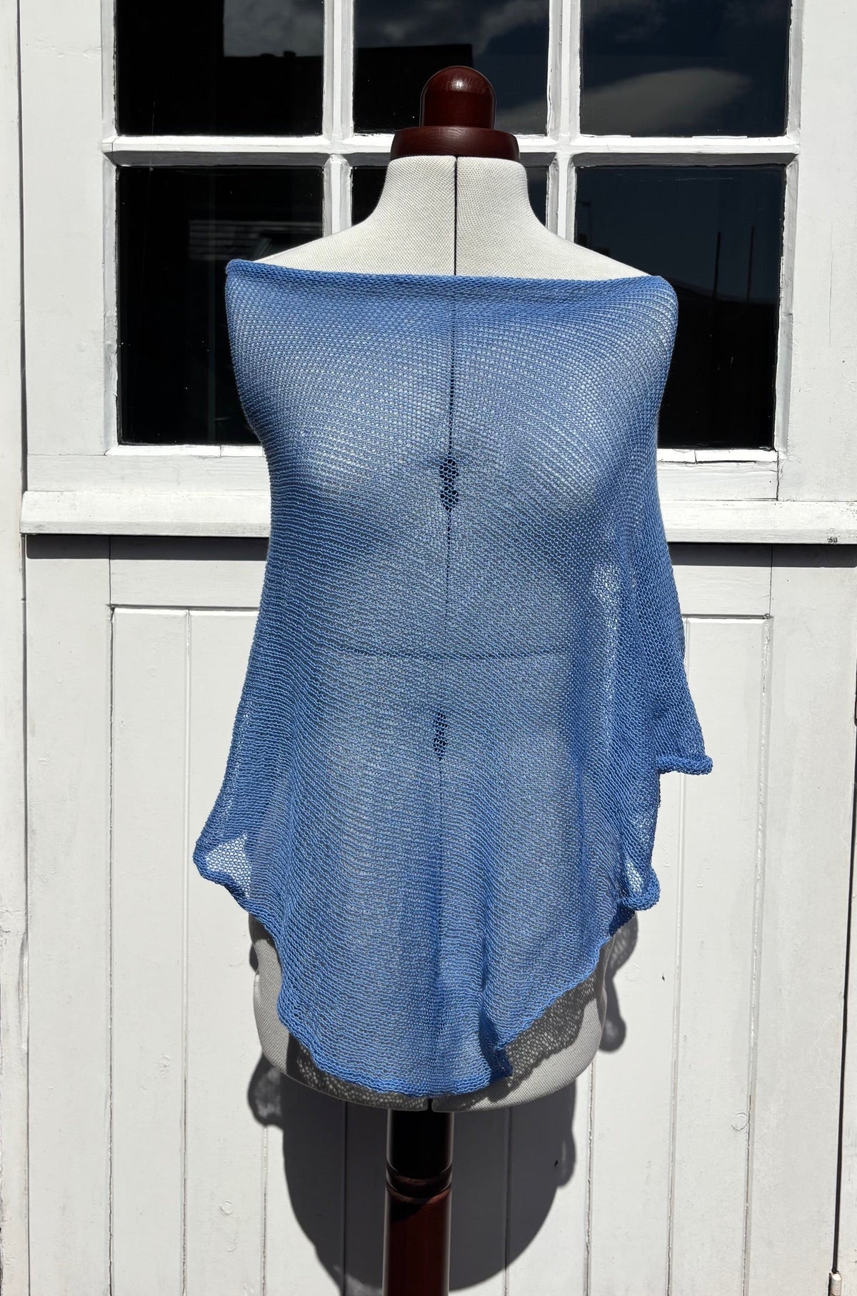A light blue open-knit shrug or poncho draped over a white dress form mannequin, set against a white wooden paneled door.