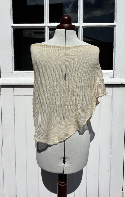 A cream-colored, semi-sheer open-knit shawl or wrap draped over a white dress form mannequin, set against a white wooden paneled wall with a window.