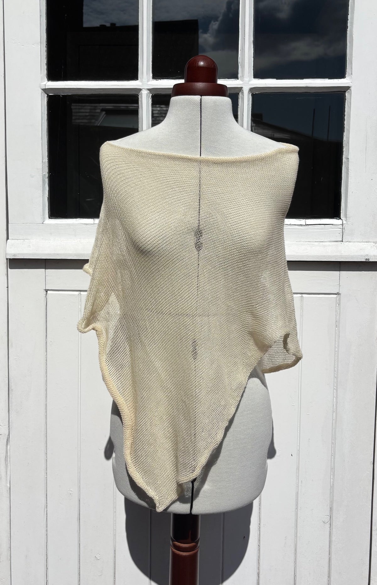A cream-colored, semi-sheer knit shawl draped over a white dress form mannequin, set against a white wooden paneled wall with a window.