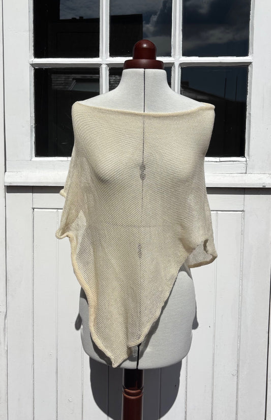 A cream-colored, semi-sheer knit shawl draped over a white dress form mannequin, set against a white wooden paneled wall with a window.