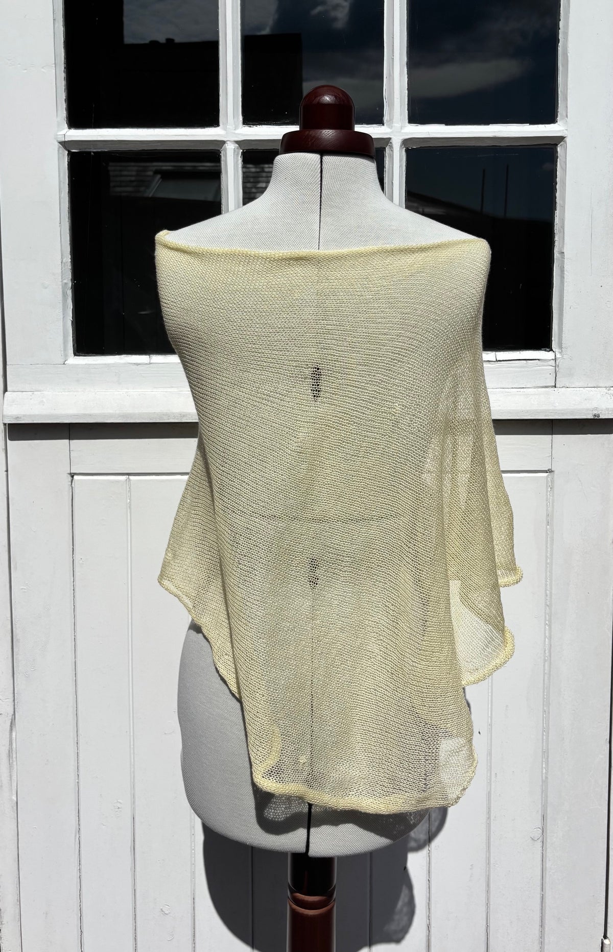 A pale yellow open-knit shawl or wrap draped over a dress form, set against a white paneled window and door background.