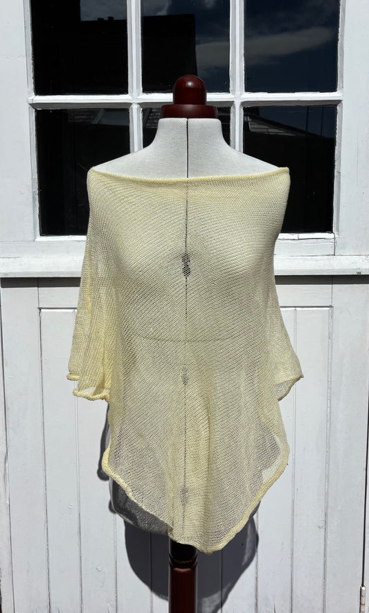 A pale yellow open-knit poncho-style top displayed on a dress form mannequin, set against a white paneled door with a window.